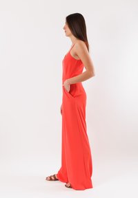 Tuta rossa realizzata in tessuto morbido, con un corpo aderente, gambe larghe e tasche laterali. Design con spalline e silhouette minimalista.