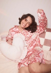 Rózsaszín mintás pizsamaszett rövid ujjú és húzózsinóros nyakkal. Egy szív alakú, fodros szegéllyel díszített párnát tart egy világos ágynemű hátterében.