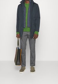 Manteau bleu marine sur un pull vert, pantalon gris, baskets olive. Tenant un sac fourre-tout noir avec des bords multicolores. Tenue décontractée en couches.