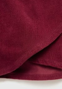 Burgundin väriinen corduroy-kangas, jossa on pehmeä rakenne ja pystysuora raihitus. Reunassa on taitettu helma viimeistellyn ilmeen saavuttamiseksi.