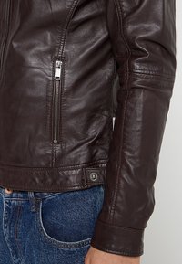 Hnědá kožená bunda s přiléhavým střihem, bočními kapsami na zip a patentkami v pase. Hladký povrch s podrobným prošitím.