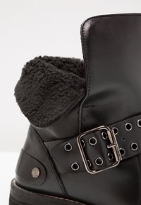 Botte en cuir noir avec une doublure en polaire douce repliée sur le dessus et une large sangle avec une boucle métallique et des œillets autour de la cheville.