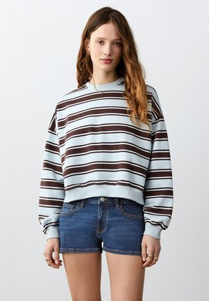 Jeune femme aux cheveux longs et ondulés portant un pull rayé bleu clair et marron ainsi qu'un short en jean, sur un fond uni.