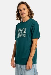 Camiseta de algodón color teal con cuello redondo, que presenta un diseño gráfico blanco en el centro. Manga corta y ajuste relajado.