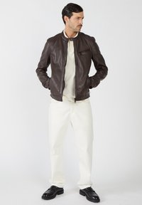 Brązowa skórzana kurtka z kołnierzem stojącym, zapinana na zamek kieszeń na piersi oraz boczne kieszenie, w połączeniu z białymi spodniami i czarnymi butami. Minimalistyczny design.