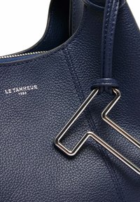 Borsa a mano in pelle blu navy con finitura testurizzata, dotata di un accento geometrico argento e chiusura con zip. Logo del marchio "LE TANNEUR" impresso.
