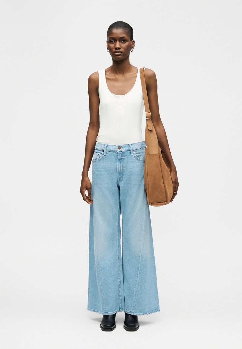 Mujer con top blanco sin mangas, vaqueros de pierna ancha azul claro, zapatos negros y lleva un bolso marrón grande al hombro, de pie sobre un fondo gris.