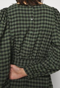 Vestido de cuadros verdes y negros con un cuello redondo, mangas largas abullonadas y un cierre de botones en la parte posterior. La tela parece texturizada y ligera.