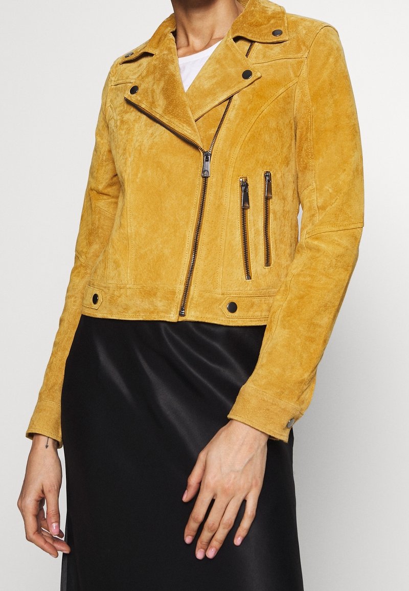 Femme portant une veste de motard en daim jaune moutarde avec des poches zippées et une jupe en satin noire, debout devant un fond uni.