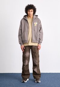 Gray zip-up hoodie with front pockets and colorful logo, beige t-shirt underneath, brown cargo pants with varied pocket sizes, and gray sneakers.
