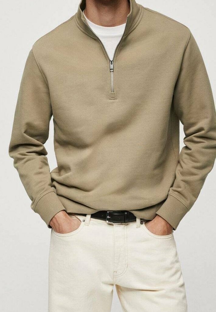 Homme portant un pull beige à fermeture éclair quart sur chemise blanche avec pantalon blanc et ceinture noire, mains dans les poches, fond neutre.