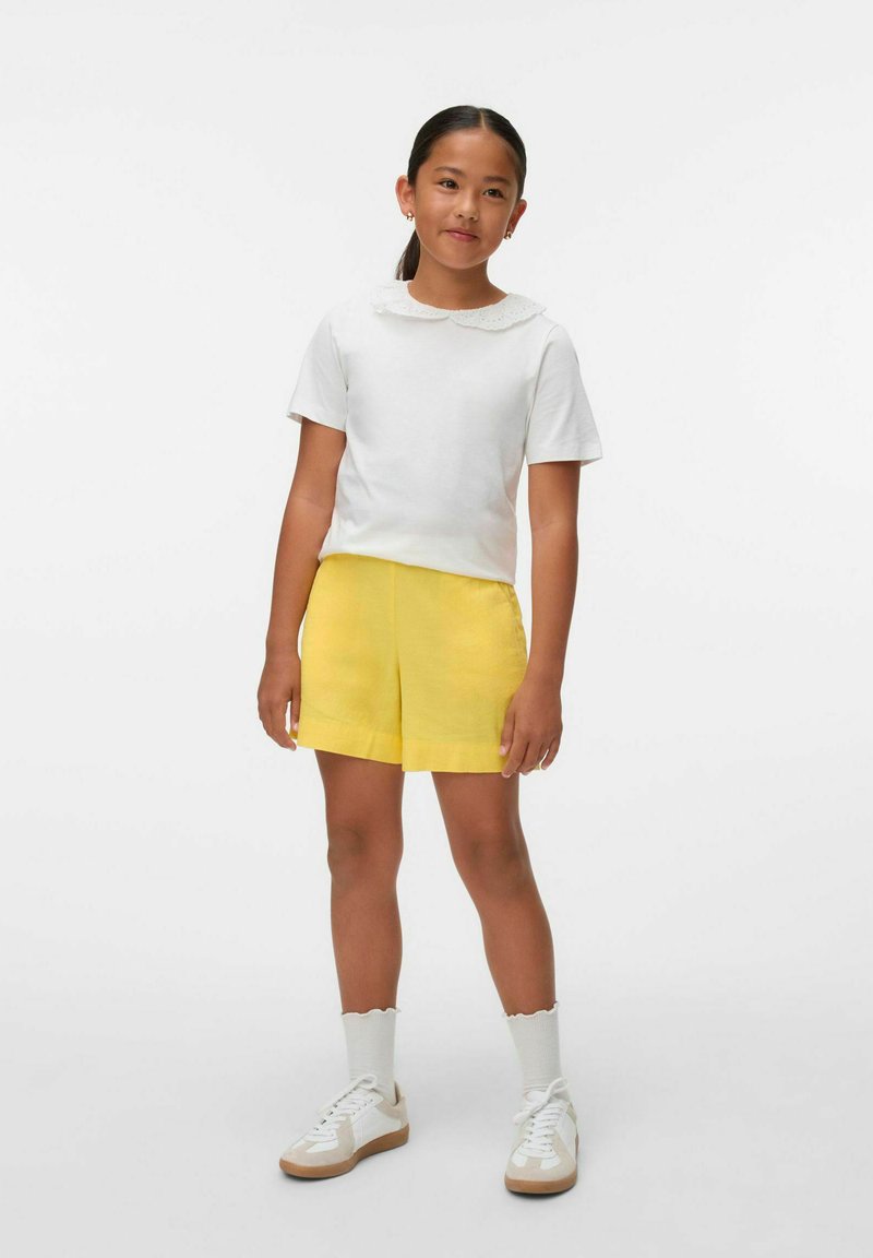 T-shirt di cotone bianco con colletto in pizzo, pantaloni corti gialli, calzini bianchi con bordi a impuntura, e sneakers bianche con accenti beige.