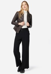 Dunkelbraune Lederjacke mit gesteppten Schulterdetails, weißer Rollkragenpullover, schwarze weite Hose und schwarze Stiefeletten.