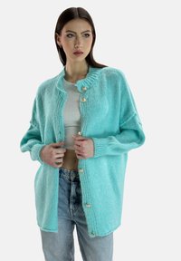 Hellblauer, gestrickter Cardigan mit einem gerippten Kragen, großen goldenen Knöpfen und oversized Passform. Über einem weißen Oberteil mit Jeans getragen.