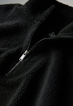 Pullover in pile nero con collo alto, mezza cerniera e superficie testurizzata. Presenta una cerniera argentata e un marchio discreto all'interno.