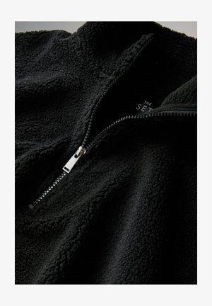 Mustast fliisist pullover, kõrge krae, pooleldi lukk ja tekstuuriga pind. Varustatud hõbedase lukuga ja peene sisesisuga.