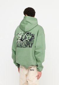 Sudadera verde de corte relajado, con una impresión gráfica en la parte posterior que incluye texto e ilustración. Puños y dobladillo de canalé.