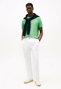 T-shirt in cotone verde con un piccolo logo, maglione in maglia blu navy annodato intorno al collo, pantaloni bianchi e sneakers beige. Design semplice e moderno.