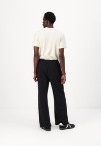 Bežna majica s kratkimi rokavi v kombinaciji s črnimi, ohlapnimi hlačami. Model nosi črne čevlje z belimi poudarki, viden od zadaj.