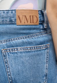 Μπλε τζιν από τζιν υλικό με καφέ δερμάτινο ταμπελάκι με την ένδειξη "VMD" στη μέση, που εμφανίζει λεπτομερή ραφή και πίσω τσέπη.