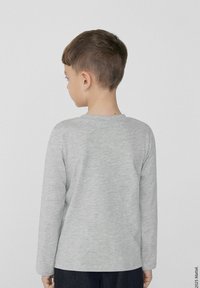 Šedá košile s dlouhým rukávem a kulatým výstřihem, vyrobená z měkkého materiálu; na dítěti stojícím zády, ukazující hladkou texturu a jednoduchý design.