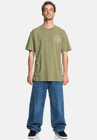 Olive green cotton t-shirt with short sleeves and a round neckline, featuring a small graphic on the chest, paired with baggy blue jeans.