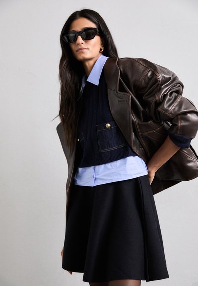Chaqueta oversized de cuero marrón, suéter de punto azul marino con bolsillos, camisa de cuello azul claro y falda negra con pliegues. Unas gafas de sol negras completan el conjunto.