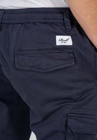 Dunkelblaue Cargo-Hose mit elastischem Bund, ausgestattet mit einer aufgesetzten Tasche und einem weißen Markenlabel auf der Rückseite. Glattes, strapazierfähiges Material.
