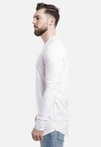 Langärmliges weißes T-Shirt mit rundem Halsausschnitt und einem Reißverschlussdetail an der Seite, getragen mit hellblauen Jeans. Glatte Stofftextur.
