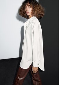 Volný bílý košile z měkké látky s klasickým límcem a zlatými knoflíkovými detaily, spárovaný s hnědými koženými kalhotami.