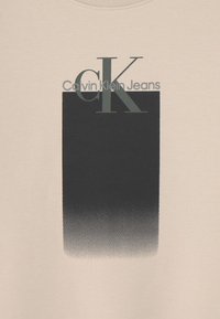 Světle béžové tričko s gradientním černým čtvercovým designem, na kterém je text "Calvin Klein Jeans" a stylizované logo "CK" v tmavě šedé barvě.