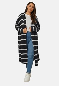 Pitkä, mustavalkoraidallinen neuletakki, jossa on huppu, avonainen etuosa, sivutaskut ja napit, yhdistettynä valkoiseen t-paitaan ja sinisiin farkkuihin.