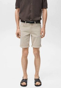 Beige, hochgekrempelte Shorts aus Denim, mit Fronttaschen und Gürtelschlaufen, kombiniert mit schwarzen Sandalen. Das Modell steht lässig.
