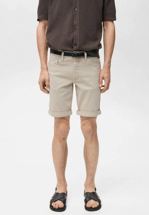 Beige, hochgekrempelte Shorts aus Denim, mit Fronttaschen und Gürtelschlaufen, kombiniert mit schwarzen Sandalen. Das Modell steht lässig.