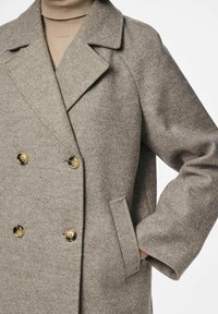 Cappotto grigio in lana doppiopetto con grandi bottoni dorati, colletto a punte, tasche laterali e tessuto texturizzato. Modello che indossa un dolcevita beige sotto.