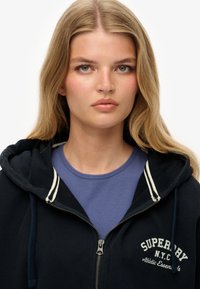 Mørkeblå zip-up hoodie med hvid stribet detalje på hætten, med et logo på brystet og båret over en lys lilla T-shirt.