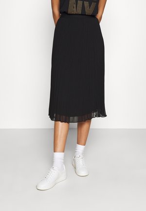 Jupe midi plissée noire avec un ourlet inférieur transparent, associée à des baskets blanches et des chaussettes mi-mollet. Le matériau semble léger et fluide.