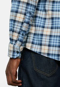 Langärmliges Hemd mit blauem Karomuster, das marineblaue und hellblaue Karos mit orangefarbenen Akzenten zeigt, Knopfmanschetten und einer Stofftextur. Über dunkler Denim getragen.