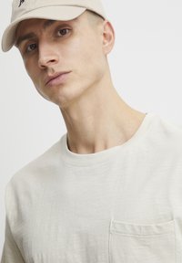 Cremefarvet kortærmet t-shirt med rund hals og brystlomme. Glat tekstur og minimalistisk design; model bærer en beige kasket.