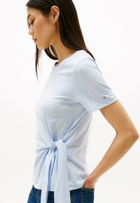 Maglietta azzurro chiaro a maniche corte con dettaglio annodato in vita. Realizzata in tessuto morbido con scollatura rotonda e logo del marchio sulla manica.