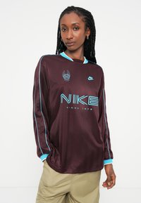 Burgundy dolgi rokavi Nike dres z svetlo modrimi poudarki in logotipom. Ima strukturirano tkanino, ovratnik in izrazit velik grafični napis "NIKE".