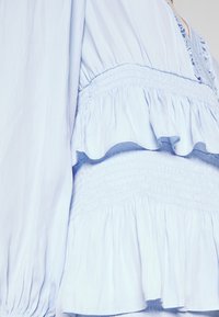 Robe bleu clair avec taille smockée, volants en étages et longue manche bouffante visible sur le côté gauche.