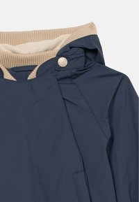 Μαρινό μπλε μπουφάν με μπεζ ριμπ λεπτομέρεια στο γιακά, διαθέτει κούμπωμα με κουμπί και λεία υφή. Ο σχεδιασμός περιλαμβάνει κουκούλα και πλαϊνές ραφές.