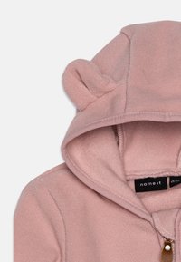 Helle rosa Fleece-Hoodie mit Ohren an der Kapuze, einem Frontreißverschluss und einer weichen, strukturierten Oberfläche. Markenlabel ist innen sichtbar.
