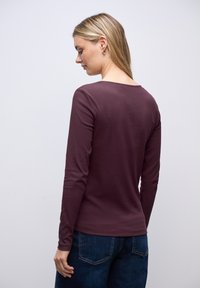 Donna con capelli biondi che indossa orecchini a cerchio dorati, un top a maniche lunghe ribbed viola e jeans blu scuri, voltata di spalle su uno sfondo neutro.