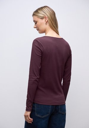 Kvinde med blondt hår iført guld hoops øreringe, en langærmet ribbet lilla top og mørkeblå jeans, vendt væk mod en ensfarvet baggrund.
