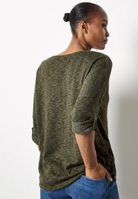 Dark green textured long-sleeve shirt with rolled cuffs, featuring side ruching and a relaxed fit, paired with blue jeans.