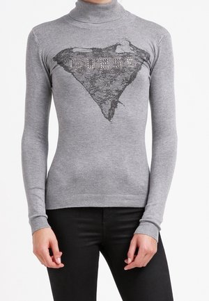 Szary dopasowany sweter z golfem z czarnym trójkątnym wzorem z koronki i zdobionym logo "GUESS" na piersi, noszony z czarnymi spodniami.