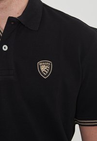 Polo shirt nero realizzato in tessuto testurizzato, con colletto, pattina con tre bottoni e un logo ricamato beige sul petto.
