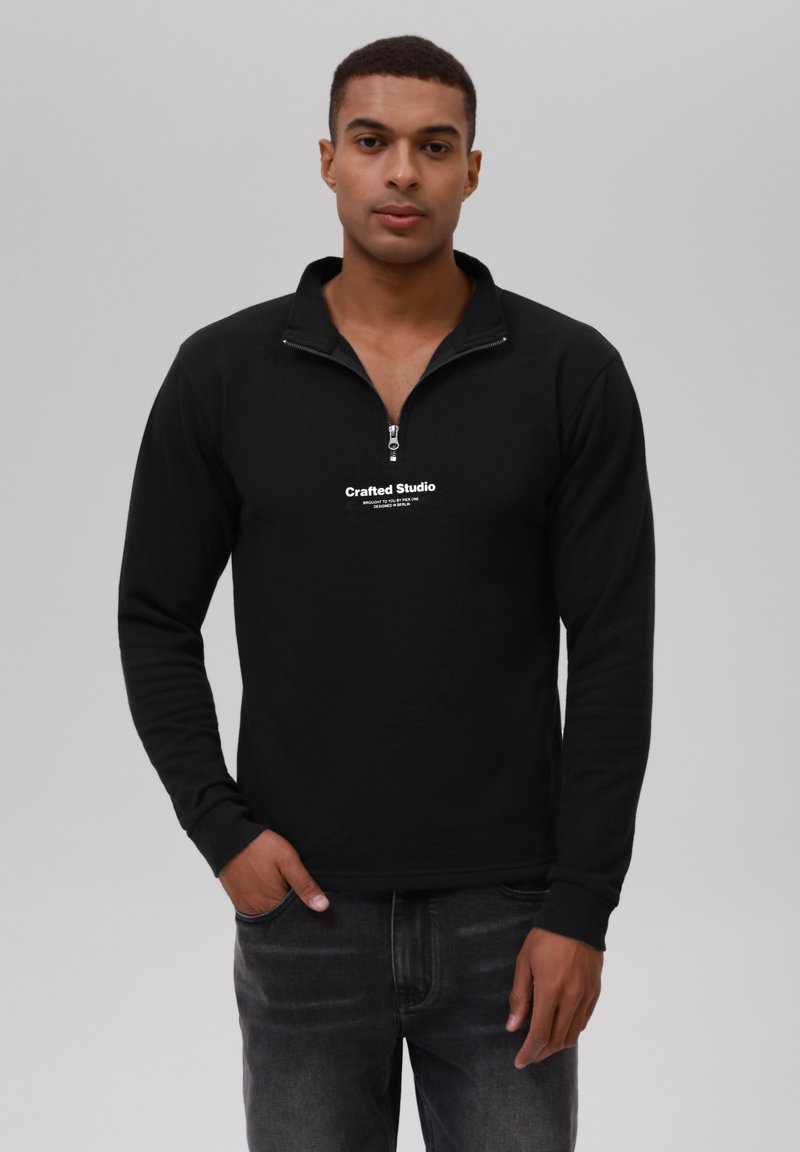 Svart zip-up hettegenser med kvart-zip nakke, med en hvit logo på brystet og ribbestrikkede mansjetter på lange ermer.
